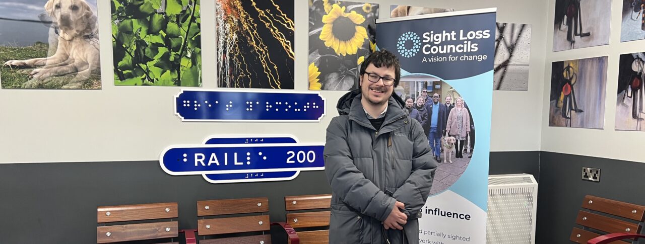 Alex, Essex SLC member, and guide dog Rosie stood in the waiting room at Great Yarmouth station, in front of the SLC Banner and exhibition artwork.