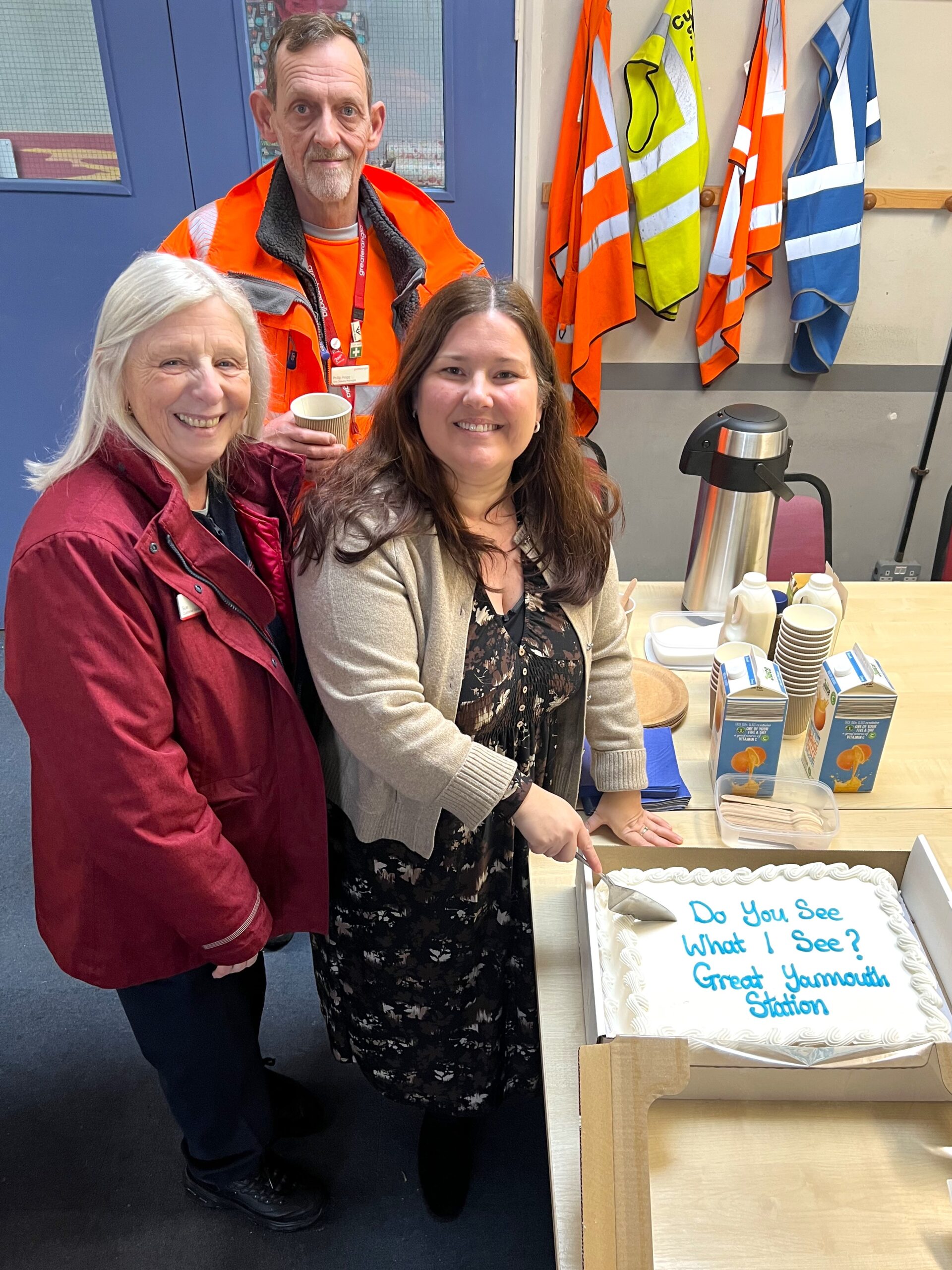 Amy Askew, Engagement Manager for East England, cutting a 'Do you see what I see?' cake. Staff members Sue and Phil are stood beside her. All are smiling at the camera.
