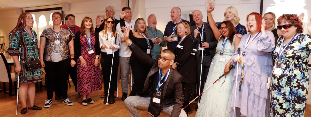 A group photo of eighteen people celebrating. They are smiling and cheering and many are wearing medals, with a few people also holding their canes. They are all smartly dressed and look happy. One man is crouched at the front with his arm in the air, celebrating.