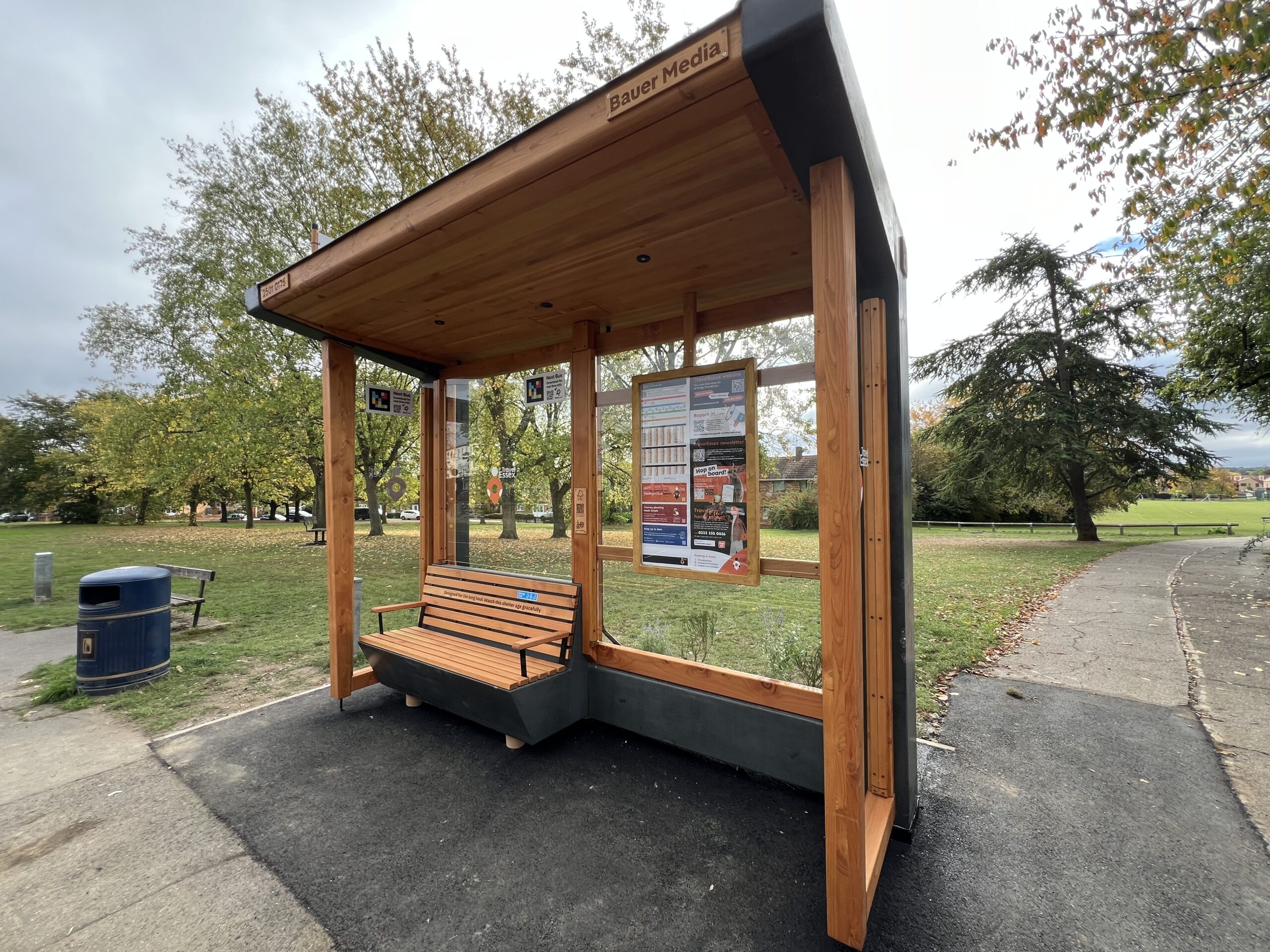 A wooden bus shelter in a park with clear panels and an angled bench designed to leave space for guide dogs. It has overhead lighting to improve visibility, large high contrast timetables, and NaviLens codes on the glass panels to support blind and low vision passengers.