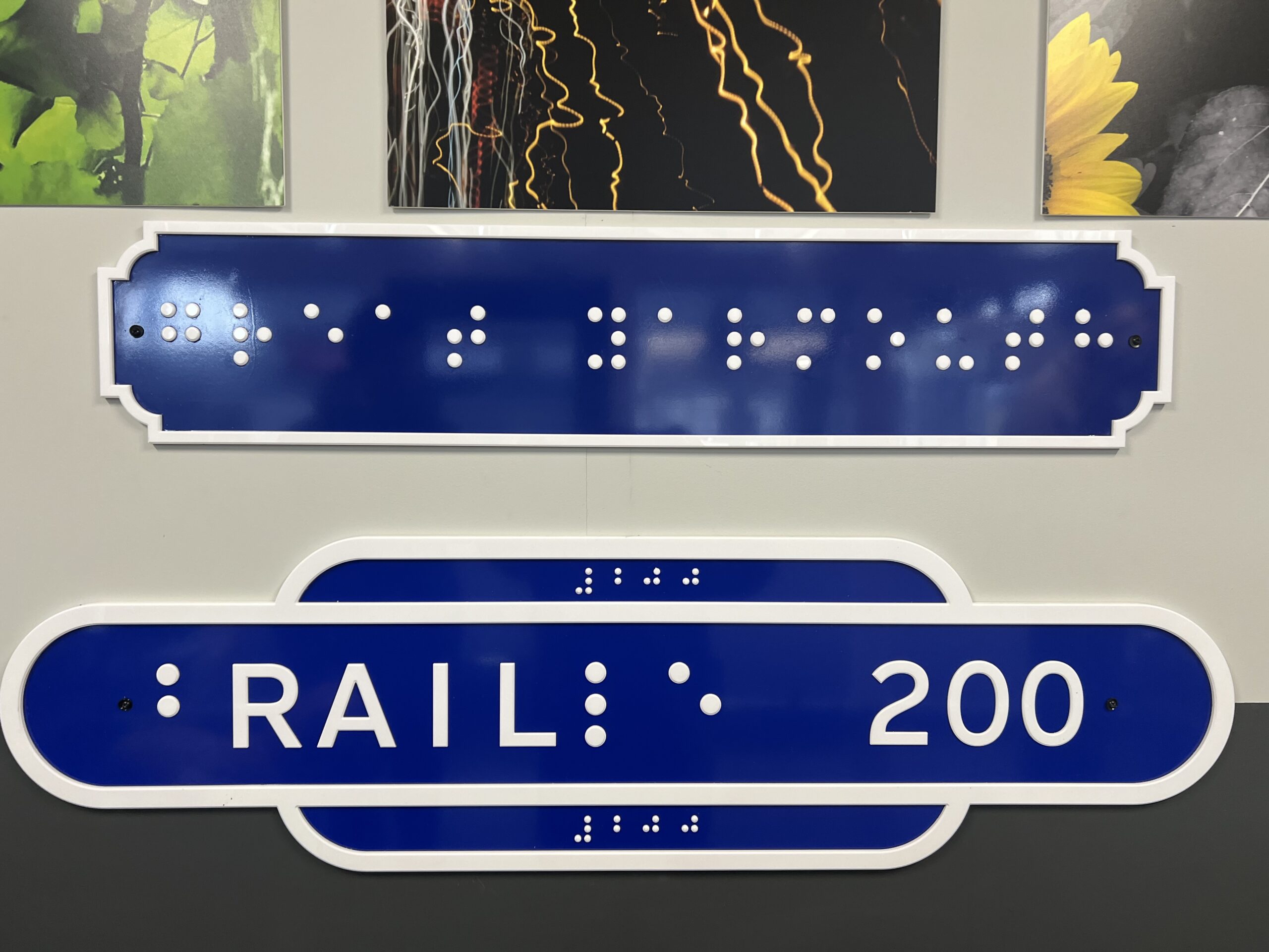 Two tactile, heritage‑style train station signs mounted on a wall. Both signs are heritage blue with white borders. The top sign features raised dots forming braille that reads “Great Yarmouth.” The lower sign displays braille for the letters b, i, and e, with the printed word “RAIL” incorporated into the braille lettering, followed by the number “200.” Braille for “200” appears at both the top and bottom edges of the lower sign. The design marks the shared 200‑year anniversaries of braille and the modern railway, both celebrated in 2025.