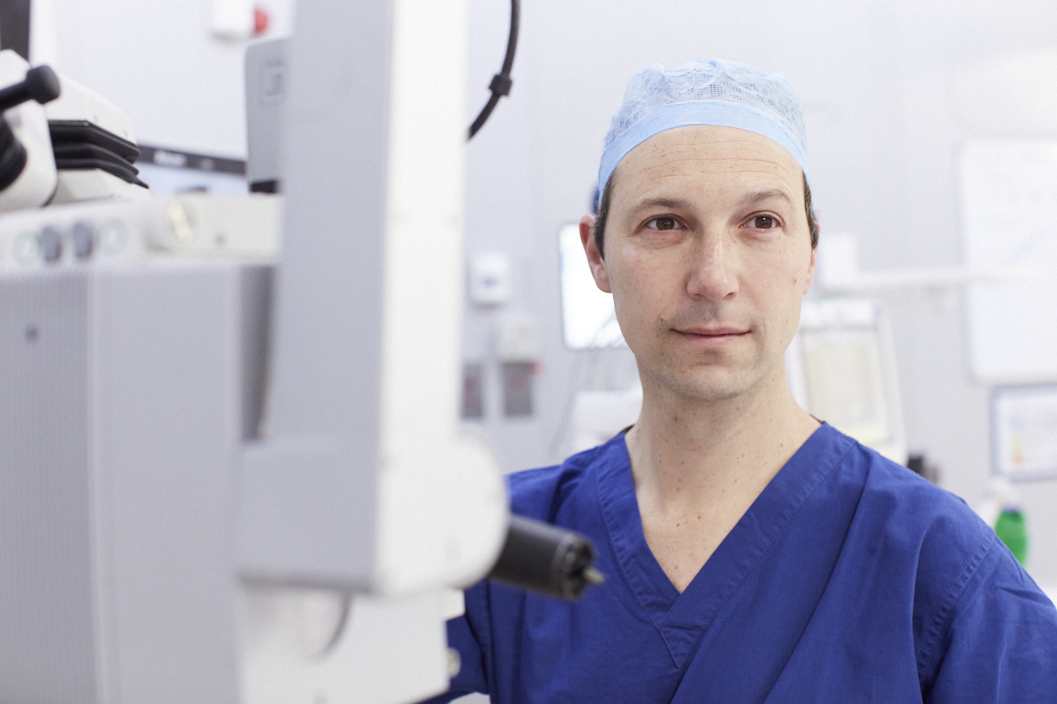 Dr Petrushking wearing blue surgical scrubs and a disposable cap is standing in clinic next to a large piece of medical equipment.