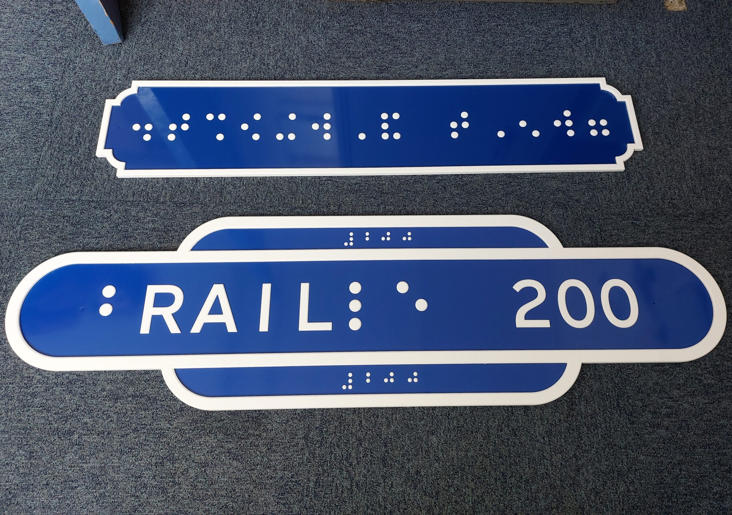 Two tactile heritage style train station signs. Both are heritage blue with white borders. The first sign features braille reading 'Great Yarmouth'. The second sign includes braille for the letters b, i, and e with the printed word 'RAIL' incorporated into the word braille. This is followed by the number 200. Braille for 200 appears at the top and bottom of the sign. The design marks the shared 200 year anniversaries of braille and the modern railway, both celebrated in 2025.
