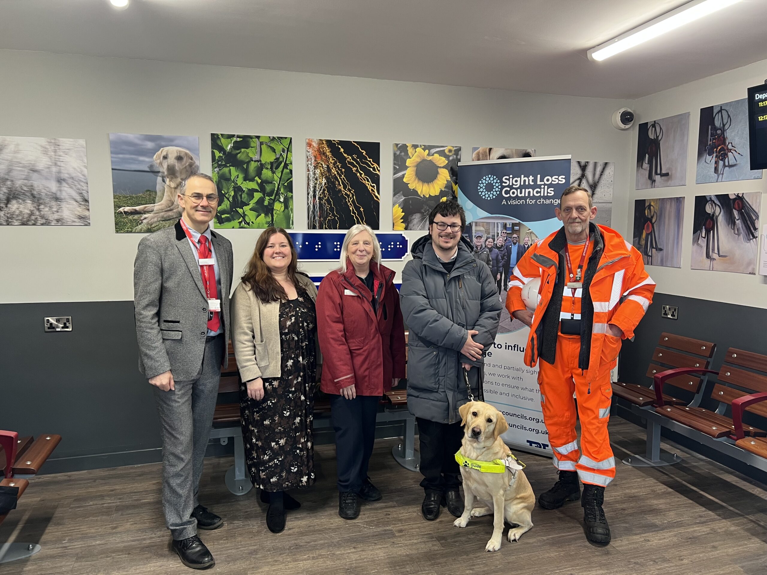 Group shot in front of the artwork at Great Yarmouth train station. From left to right: Scott, Greater Anglia Partnerships Manager, Amy, Engagement Manager for East England, Sue, Customer service at Greater Anglia, Essex SLC member Alex and guide dog Rosie, and Phil, Site Manager for Greater Anglia.
