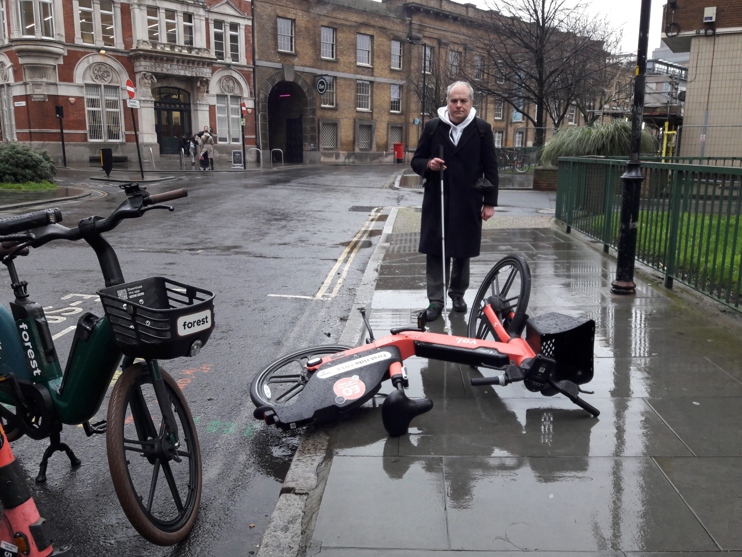 Liam O'Carroll, Interim Engagement Manager for London Sight Loss Councils, is walking along a wet pavement with his long cane. In his path, is a fallen over e-bike, blocking the way,