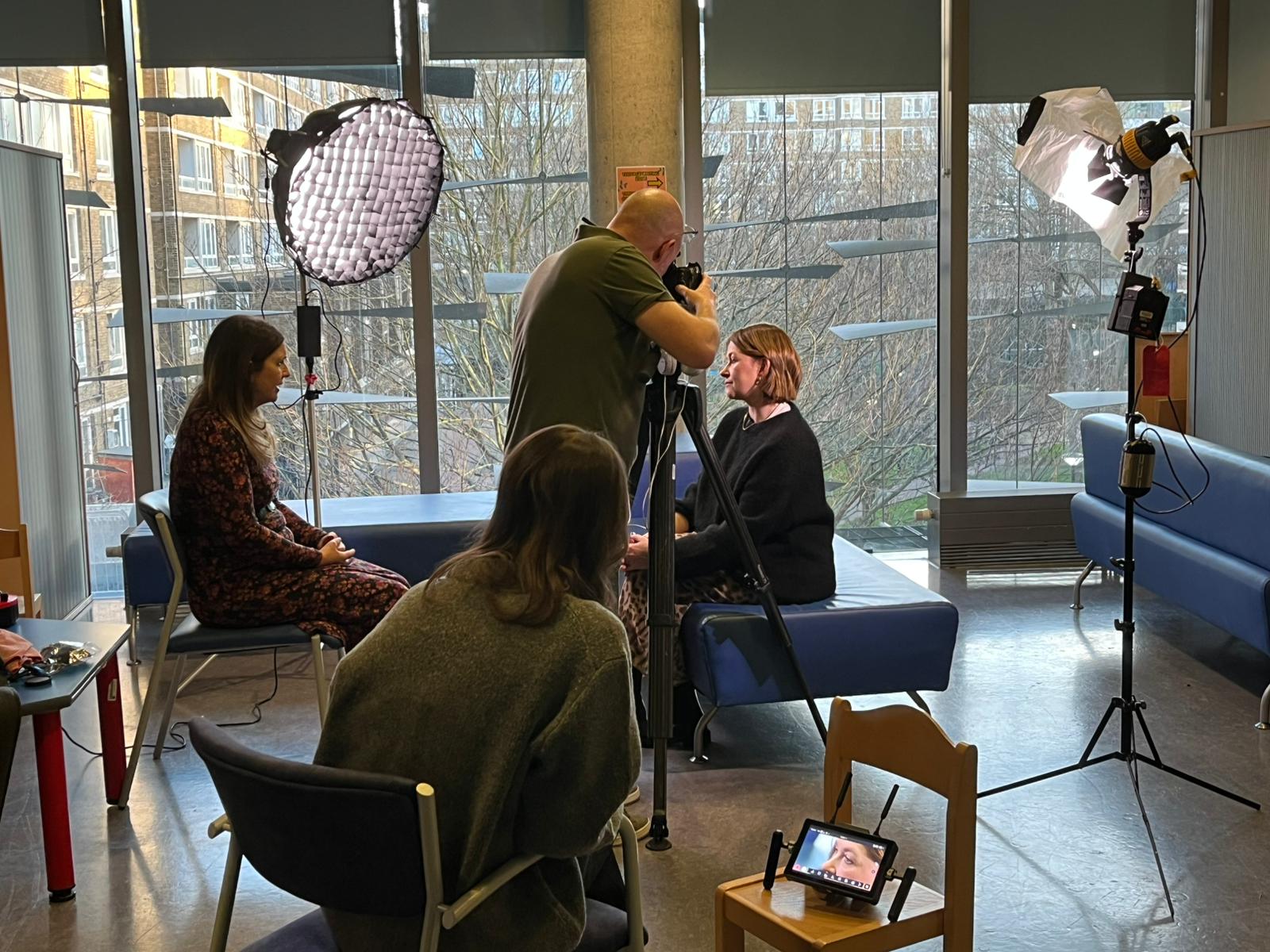A behind the scenes view of Nicki being interviewed by the BBC at Moorfields. Two large studio lights with diffusers are positioned of either side of Nicki and reporter, Sophie Hutchinson. A cameraman is standing in front of Nicki, filming her, whilst another is seated in the foreground, watching a small monitor.