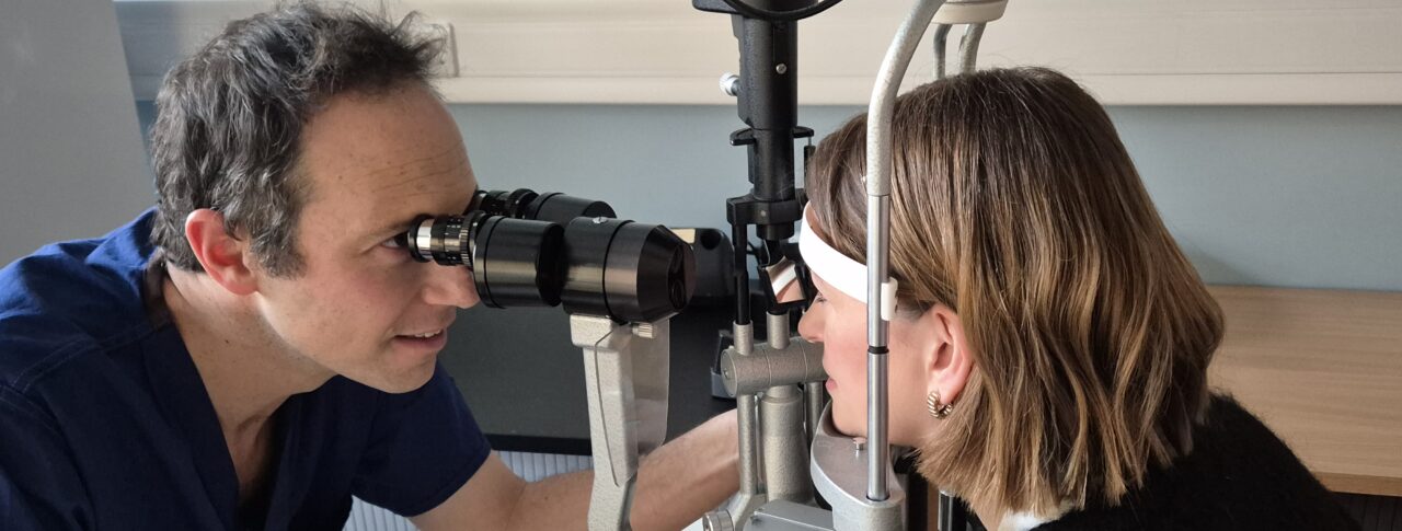 Dr Petrushkin examines Nicki's eyes using a slit lamp in clinic. Dr Petrushkin is seated on the left, wearing dark blue scrubs, whilst Nicki is positioned on the right with her chin on the rest, leaning forward towards the device.