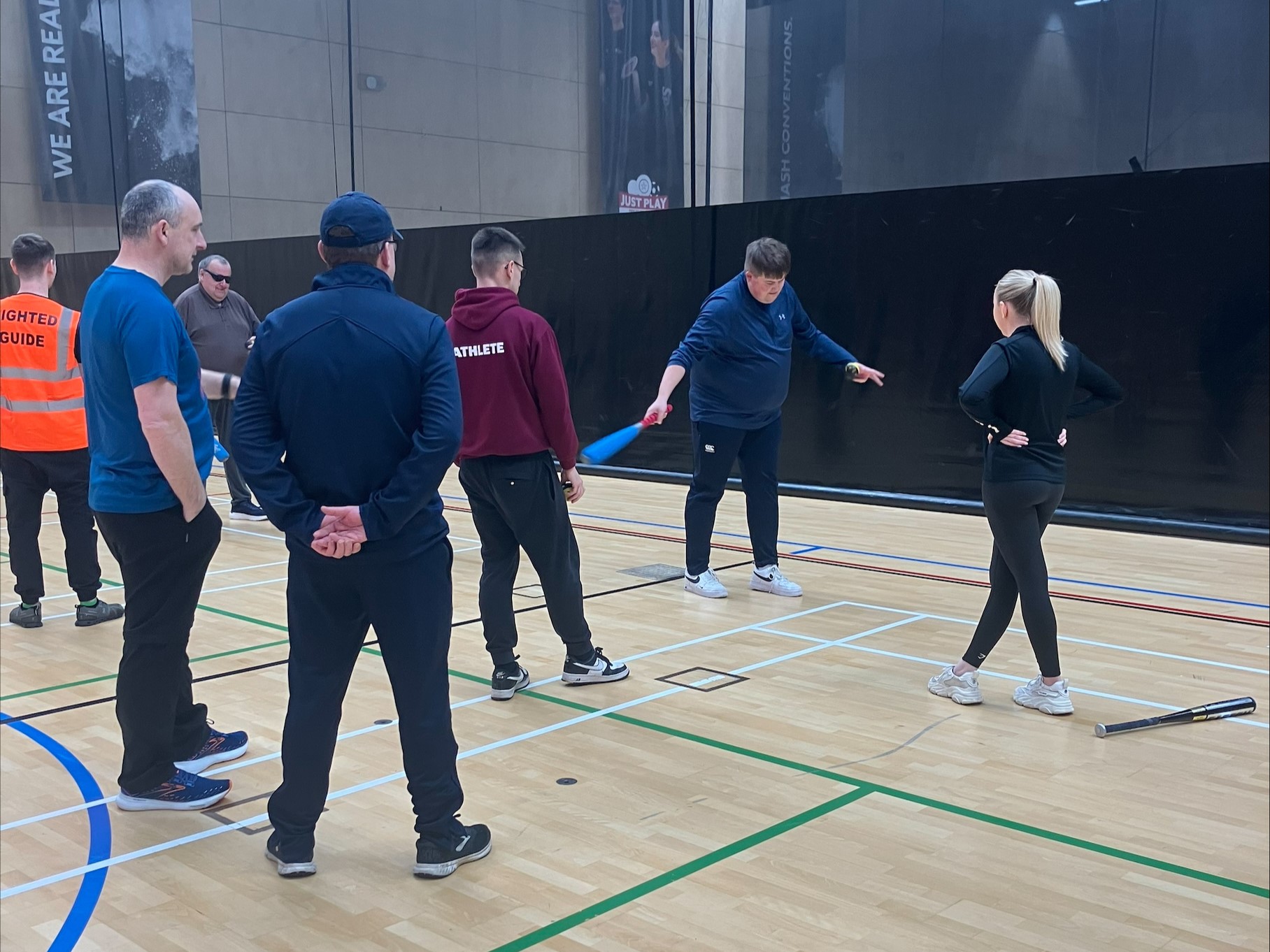 An indoor sports session at the North East Let's Get Active Day. A cricket coach is demonstrating a movement with a bat whilst attendees watch and listen to his instruction.