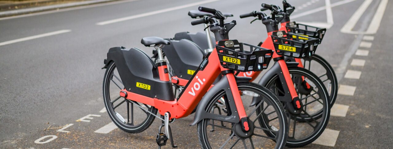 Three red and black Voi rental e‑bikes are parked neatly in a designated bike bay alongside a city street. They are lined up facing forward, each with a front basket displaying a yellow ID tag. Cars and buildings are visible in the background on the opposite side of the road.