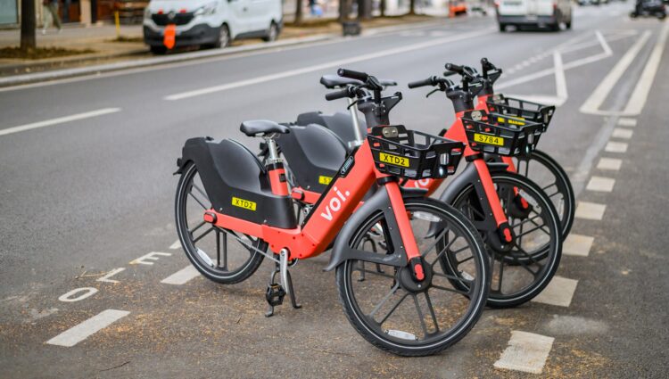 Three red and black Voi rental e‑bikes are parked neatly in a designated bike bay alongside a city street. They are lined up facing forward, each with a front basket displaying a yellow ID tag. Cars and buildings are visible in the background on the opposite side of the road.