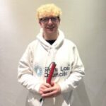 Photo of Antony, Greater Manchester SLC member. Antony has blond, curly, hair and is wearing glasses. He is stood against a white wall and wearing a white Sight Loss Councils hoodie. He is holding his long cane, smiling at the camera.