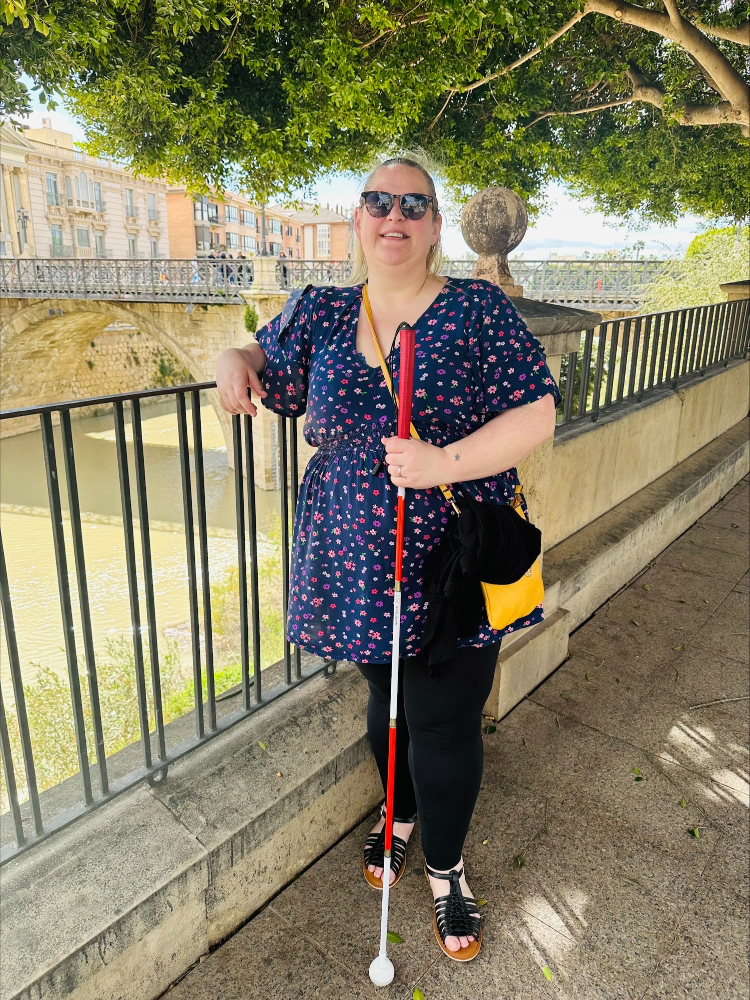Dannie, Derbyshire Sight Loss Council member, stands outdoors beside a metal railing overlooking a river and an old stone bridge. She holds a long white cane with red stripes, indicating dual -sensory loss. She is wearing a navy blue dress with a pink floral pattern, black leggings, and sandals, with a yellow bag at her side. Overhead, leafy trees cast dappled shade, and sunlit buildings line the riverbank in the background.
