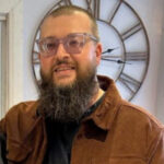Photo of Neil, Merseyside SLC member. Neil has a long, full beard and is standing indoors, in front of a large decorative wall clock, He is wearing a brown, corduroy overshirt over a black t-shirt. He is wearing glasses with a clear frame, and smiling at the camera.