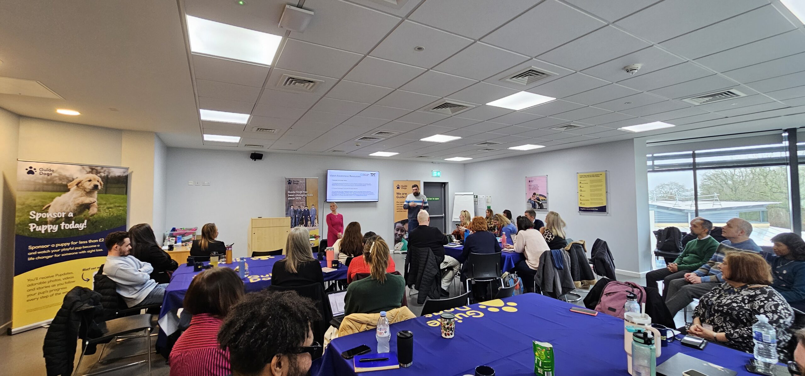 Large group of people seated around tables in a bright training room, listening to Jack and Kelly, Engagement Managers for Sight Loss Councils, presenting at the front, near a screen. The tables are covered in blue cloths with Guide Dogs' logo, and posters about guide dogs and puppy sponsorship are displayed around the room.