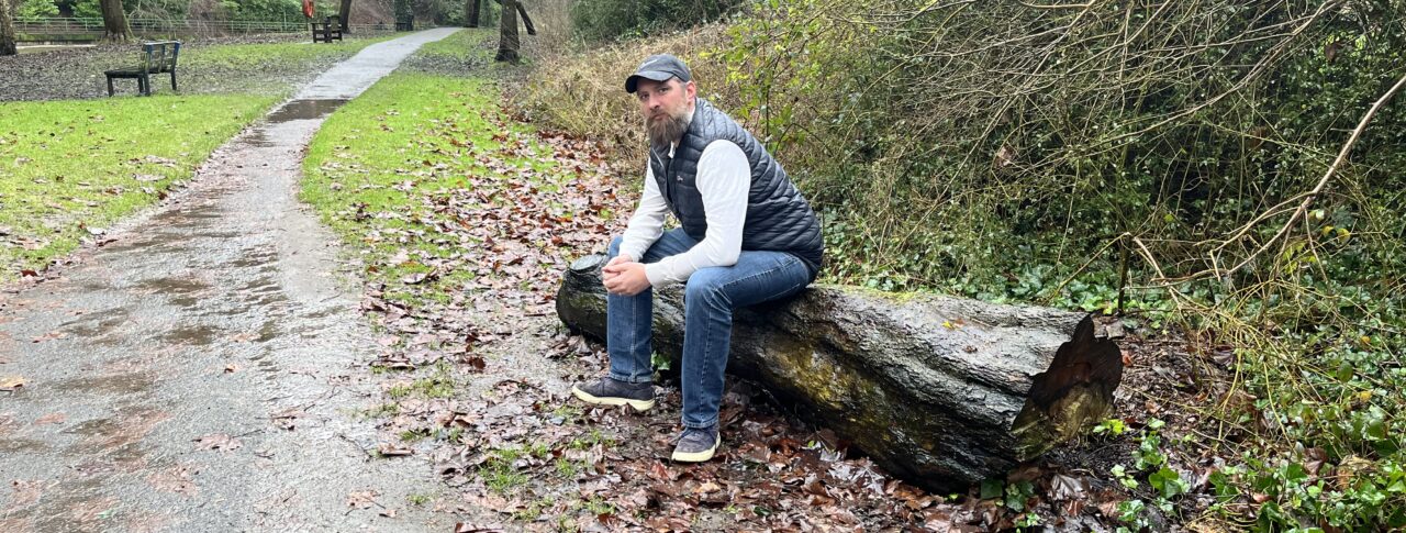 Jack Moffat, Engagement Manager for North East England, is sat on a large, fallen, tree trunk to the side of a wet, muddy, path.