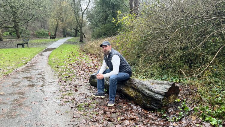 Jack Moffat, Engagement Manager for North East England, is sat on a large, fallen, tree trunk to the side of a wet, muddy, path.