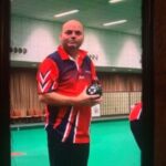 Photo of John, Bedfordshire and Hertfordshire SLC member. He is wearing a red, navy, and white sports shirt and is standing on an indoor bowls green, holding a black bowls ball in both hands. The photo appears to be taken through a window, with dark curtains or a frame, framing the shot.
