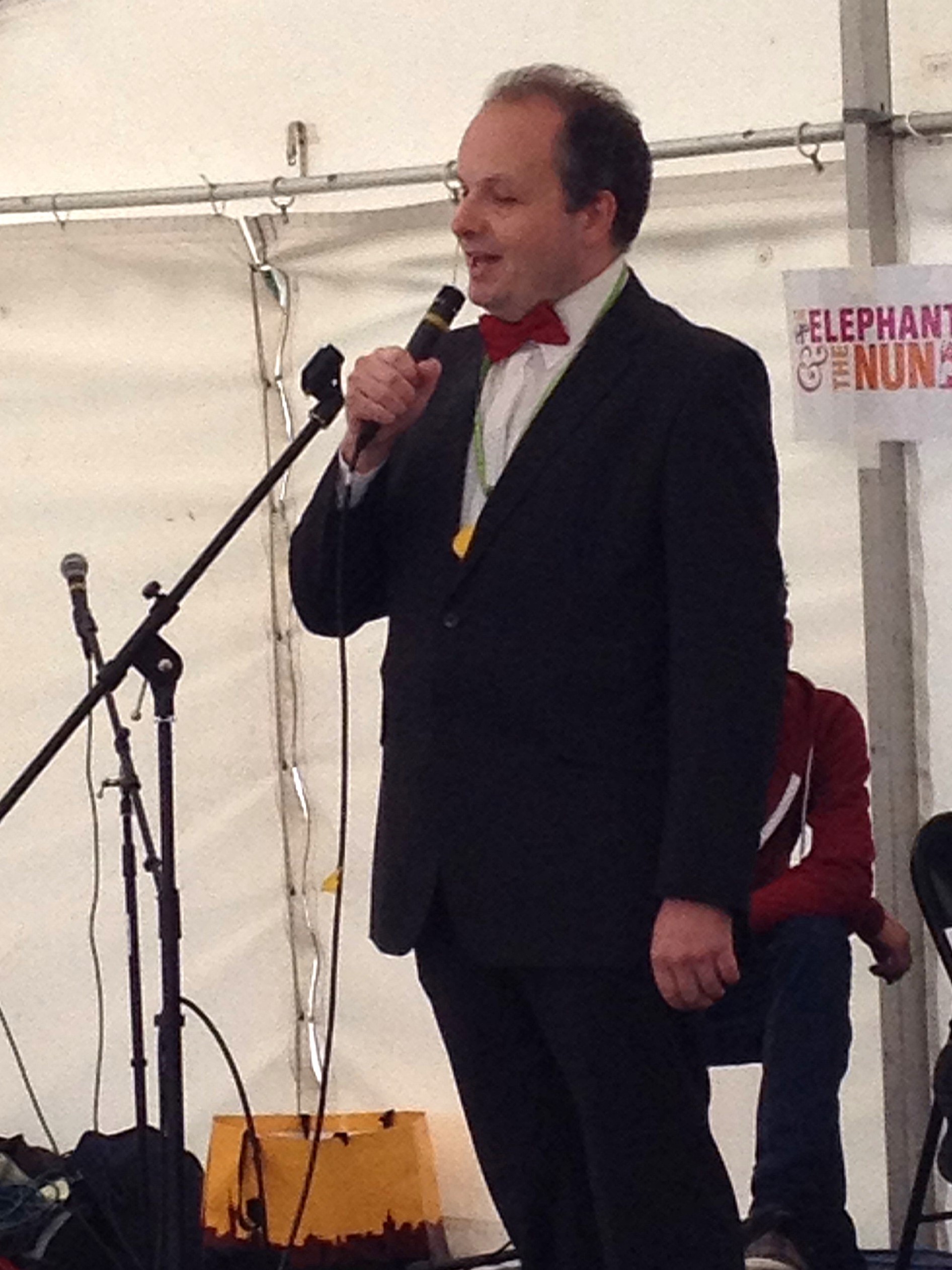 Liam O'Carroll, Engagement Manager for London SLCs is stood on stage in a white tent, holding a microphone. He is wearing a dark suite and burgundy bow tie. A seated person is visible in the background, next to a sign which reads 'Elephant & the Nun'.