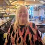Photo of Paula, Bedfordshire and Hertfordshire SLC member. Paula is standing in a restaurant with a pitched roof and exposed beams - set tables are in the background. She has long, blonde hair and is wearing a pink and black print top. She is smiling at the camera.