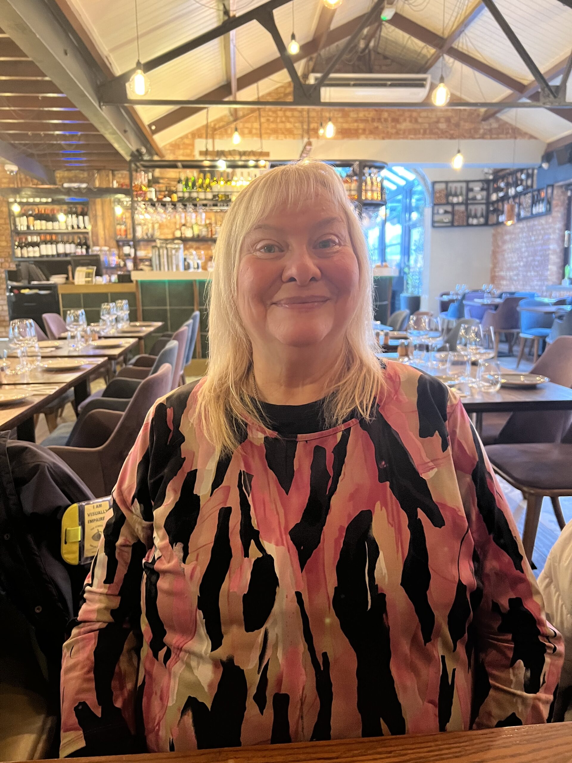Photo of Paula, Bedfordshire and Hertfordshire SLC member. Paula is standing in a restaurant with a pitched roof and exposed beams - set tables are in the background. She has long, blonde hair and is wearing a pink and black print top. She is smiling at the camera.