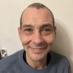 Headshot of Stuart, Bedfordshire and Hertfordshire SLC member. Stuart is standing in from of a cream wall, smiling at the camera. He has short dark hair, and a blue/grey jumper on.