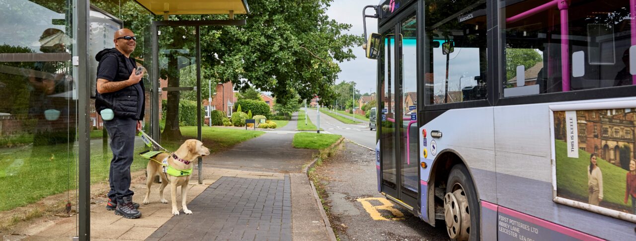 A man, smiling and holding a mobile phone, is standing at a bus stop with his guide dog wearing a harness, waiting as a bus pulls up to the curb. The scene is outdoors on a residential street with greenery and houses in the background.