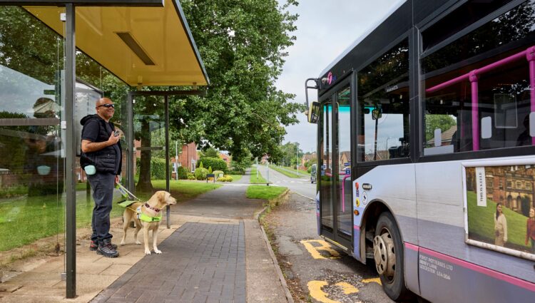 A man, smiling and holding a mobile phone, is standing at a bus stop with his guide dog wearing a harness, waiting as a bus pulls up to the curb. The scene is outdoors on a residential street with greenery and houses in the background.