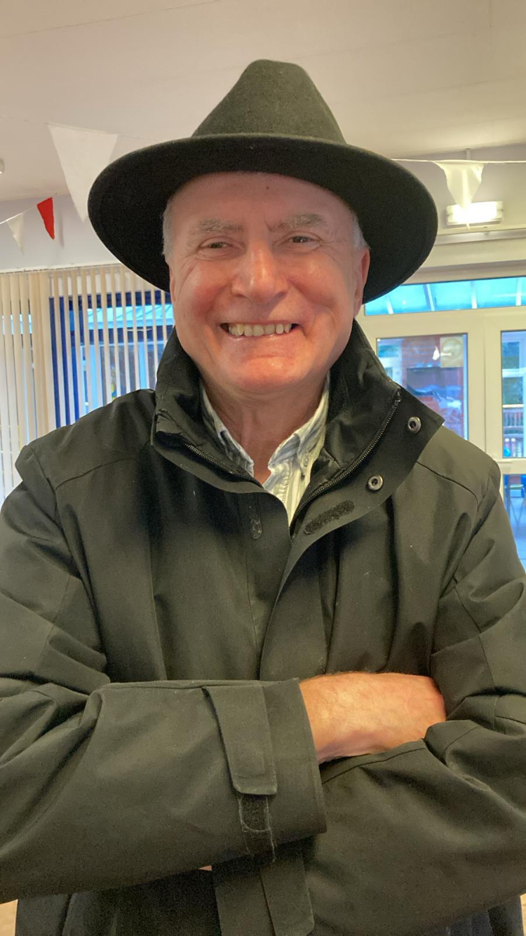 Photo of Johnny, Lancashire SLC member. He is wearing a black wide‑brimmed hat and a dark coat and stands indoors with his arms crossed. Behind them is a bright room with large windows, vertical blinds, and string decorations hanging from the ceiling.