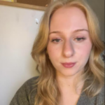 Selfie of Amna, Birmingham and Black Country SLC member. She has long, light-coloured hair, and is wearing a grey top. She is standing indoor, smiling at the camera.