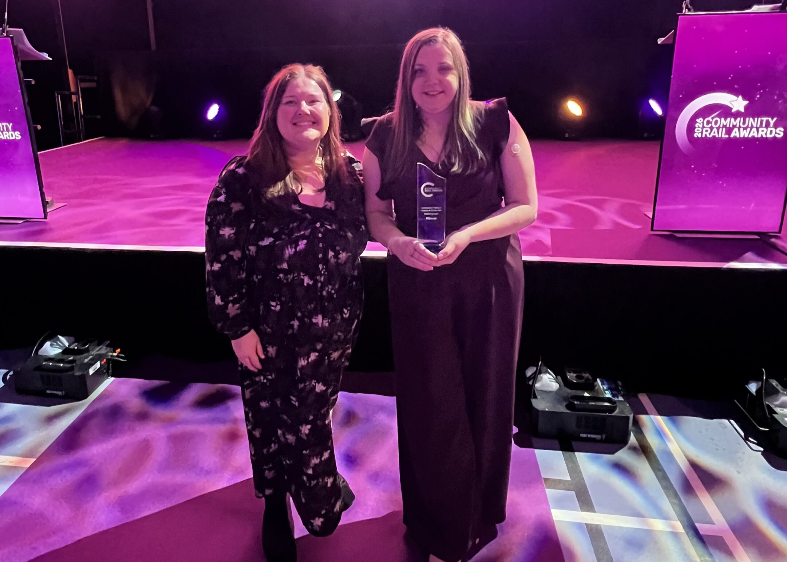 Samantha Leftwich, Engagement Manager for East England, and Amy Askew, Engagement Manager for East England (Maternity Cover) are standing next to the stage. Sam is holding a glass award. The background is lit with purple and pink lighting, with a Community Rail Awards podium either side of the stage. Both Amy and Sam are wearing full-length, black dresses.