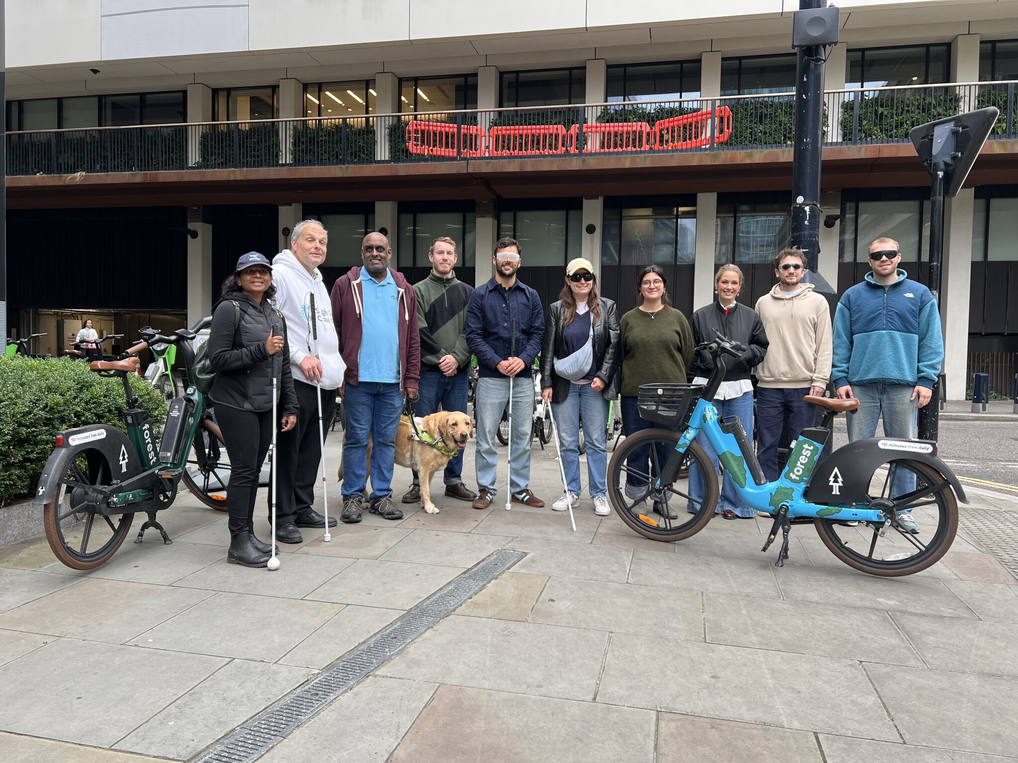 A group shot of Forest staff and London SLC members following our sim-spec walk. Everyone is lined up, on the pavement, smiling at the camera. There are Forest bikes scattered around the group and staff are wearing simulation glasses.