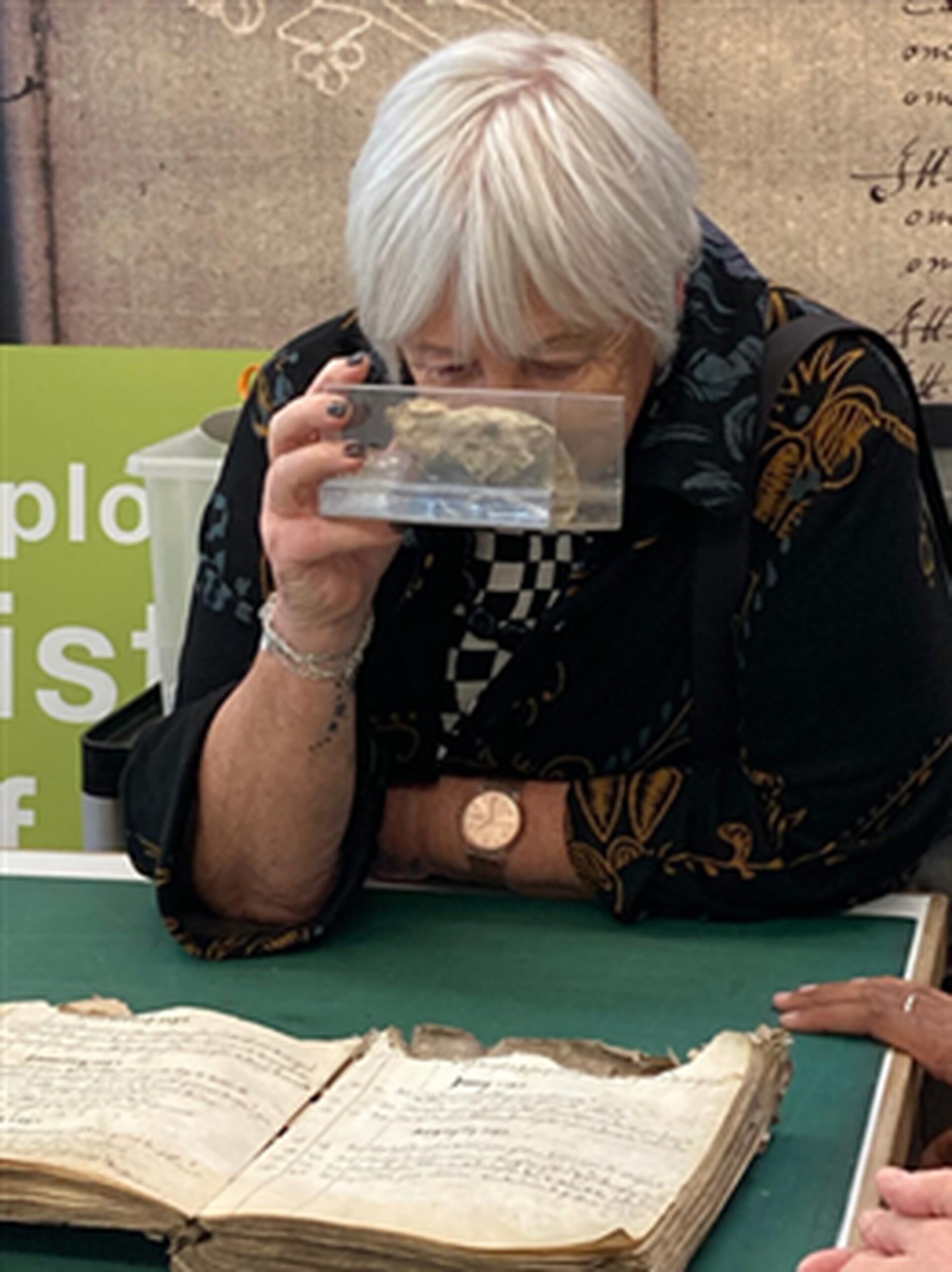 Ann, Gloucestershire SLC member, is seated at a table whilst examining an artefact in a glass case. She is holding it up to her face. Beneath her, is an old, open, book, filled with dense, handwritten script.