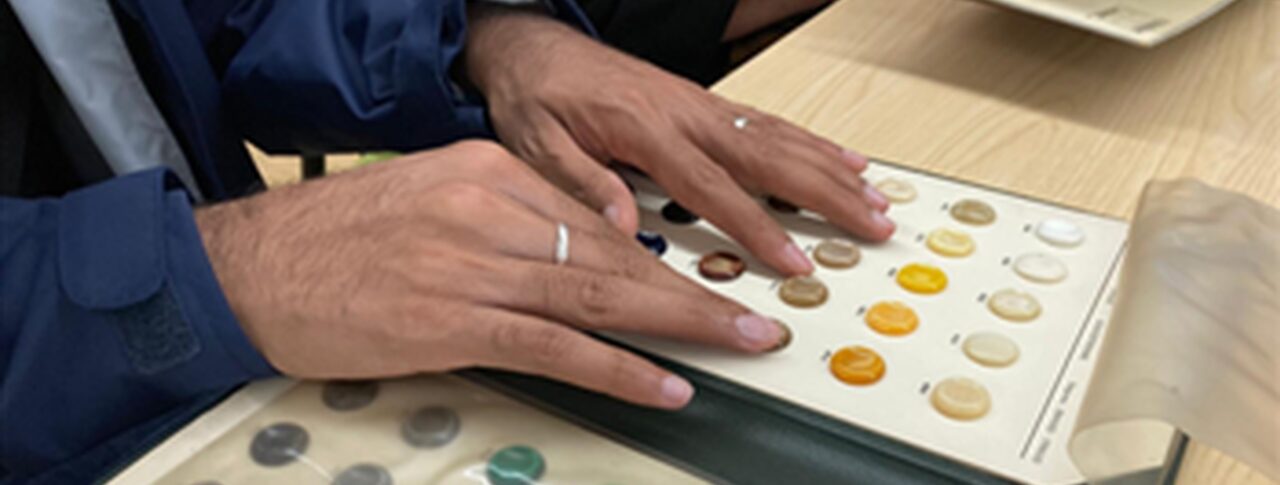 A close-up of a person's hands as they run their fingers over a display book of raised, circular samples in various colours. A second person is beside them, looking through a similar book.