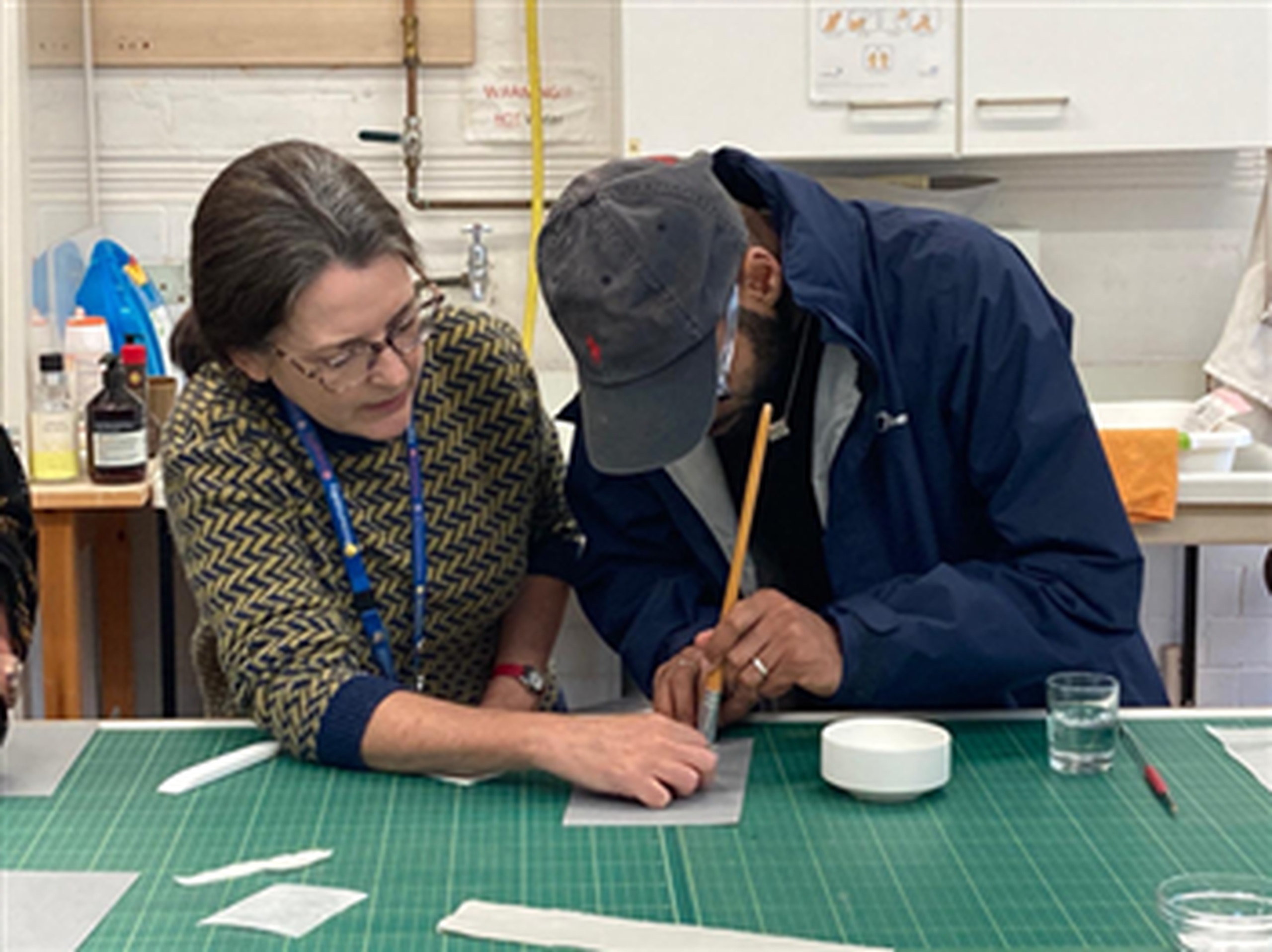 Yahya and a female document restorer are working together at a large cutting mat table. Yahya's hand is being guided as they use a long wooden tool to mark or measure a piece of material on the table. Various tools and supplies, including a small bowl, a glass of water, and sheets of material, are spread out around them. Shelves, bottles, and equipment are visible in the background.