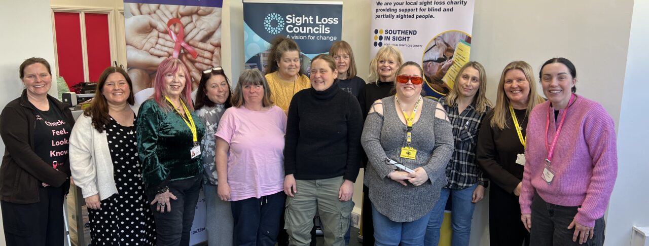 A group photo following Essex SLC's breast cancer screening event. Blind and partially sighted attendees are stood alongside Amy Askew, Engagement Manager for Thomas Pocklington Trust, Angela and Lauren from Lady McAdden, and Lucy from Southend in Sight. They are stood facing the camera, smiling.