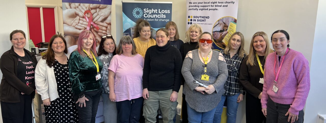 A group photo following Essex SLC's breast cancer screening event. Blind and partially sighted attendees are stood alongside Amy Askew, Engagement Manager for Thomas Pocklington Trust, Angela and Lauren from Lady McAdden, and Lucy from Southend in Sight. They are stood facing the camera, smiling.