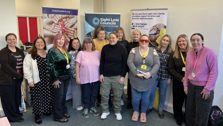 A group photo following Essex SLC's breast cancer screening event. Blind and partially sighted attendees are stood alongside Amy Askew, Engagement Manager for Thomas Pocklington Trust, Angela and Lauren from Lady McAdden, and Lucy from Southend in Sight. They are stood facing the camera, smiling.