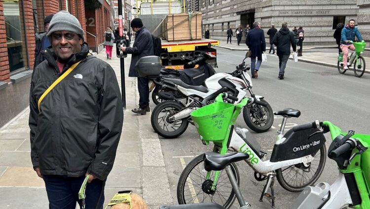 SW London member, Haren, is standing next three Lime bikes with one parked over the curb and protruding onto the path. Haren is with his guide dog, Addy.