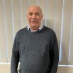 Photo of Geoff, Staffordshire SLC member. Geoff is standing indoors in front of a set of white, vertical blinds. He is wearing a dark grey jumper over a light coloured shirt.