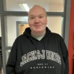 Photo of Ian, Staffordshire SLC member. He is wearing a black Jack & Jones hoodie and is standing indoors, in front of a grey-framed window and red wall. Ian is smiling at the camera.