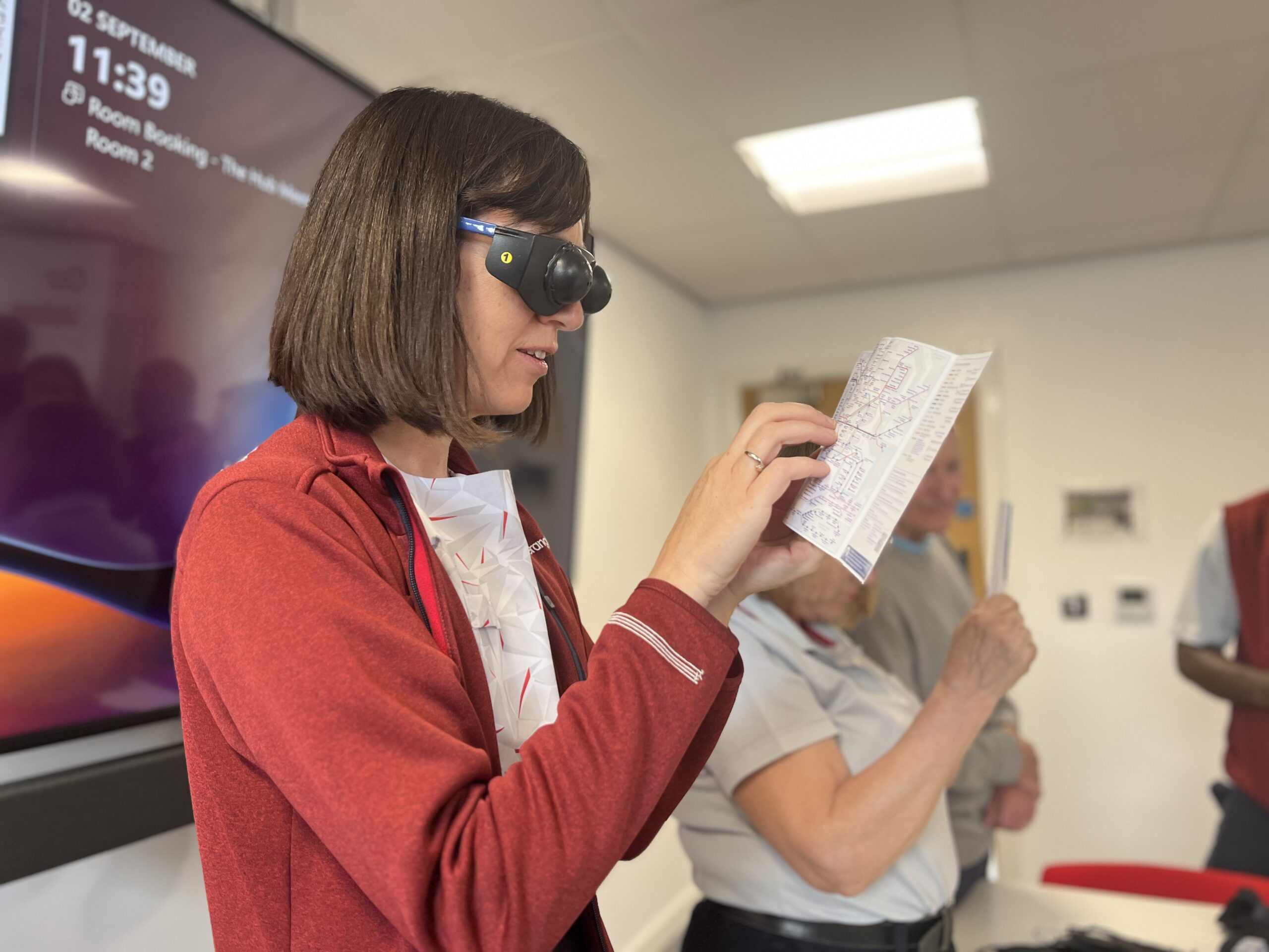 Kat, Greater Anglia staff member, is pictured with a pair of simulation glasses on and trying to read a travel leaflet. which she is holding up in front of her face.