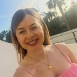 Selfie photo of Katharine, SW London SLC member. She is standing outdoors in a pink, strappy top and a gold necklace. She has long, brown hair. Behind her are palm trees, a white wall, and soft sunlight in the sky.