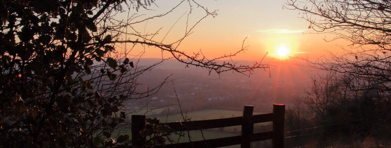 A scenic sunset view from Box Hill in Surrey. The sun is low on the horizon, casting a warm orange glow across the sky and landscape below. Tree branches frame the photo on both sides, and a wooden fence stands in the foreground, overlooking the valley. In the distance, hills and fields appear under the fading daylight.