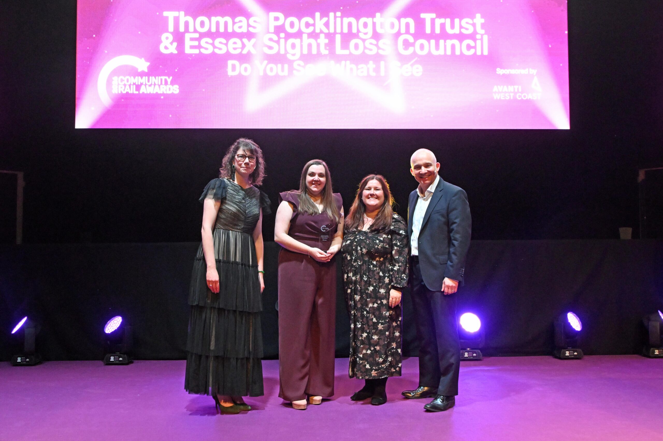 Samantha Leftwich and Amy Askew, Engagement Managers for East England, stand on stage smiling with the presenters after being announced as winners at the Community Rail Awards 2026. Behind them is a screen displaying “Thomas Pocklington Trust and Essex Sight Loss Council – ‘Do You See What I See?’” and the Community Rail Awards logo.