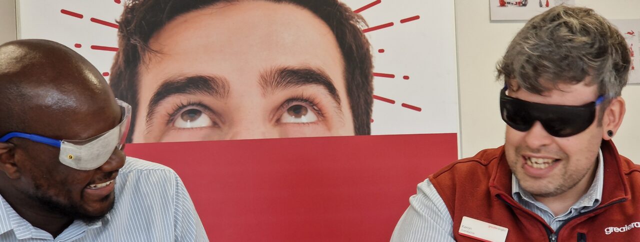 Two male staff members sit at a table, engaged in an activity during the VI Awareness session. Both are wearing simulation glasses and looking towards each other. The person on the right is wearing a light blue, short sleeved Greater Anglia shirt and the person on the right is wearing a red sleeveless vest over his light blue shirt. Behind them, a large red and white Greater Anglia display board features large text and graphics.