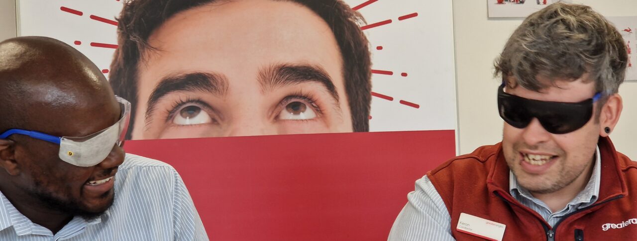 Two male staff members sit at a table, engaged in an activity during the VI Awareness session. Both are wearing simulation glasses and looking towards each other. The person on the right is wearing a light blue, short sleeved Greater Anglia shirt and the person on the right is wearing a red sleeveless vest over his light blue shirt. Behind them, a large red and white Greater Anglia display board features large text and graphics.