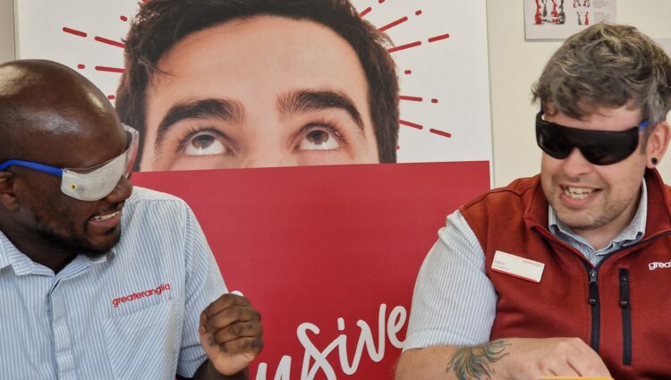 Two male staff members sit at a table, engaged in an activity during the VI Awareness session. Both are wearing simulation glasses and looking towards each other. The person on the right is wearing a light blue, short sleeved Greater Anglia shirt and the person on the right is wearing a red sleeveless vest over his light blue shirt. Behind them, a large red and white Greater Anglia display board features large text and graphics.