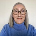 A headshot of Zeta, Tyne and Wear SLC member. She has light grey, shoulder-length hair, and is wearing glasses with dark frames and a blue roll-neck sweater. She is facing the camera, smiling.