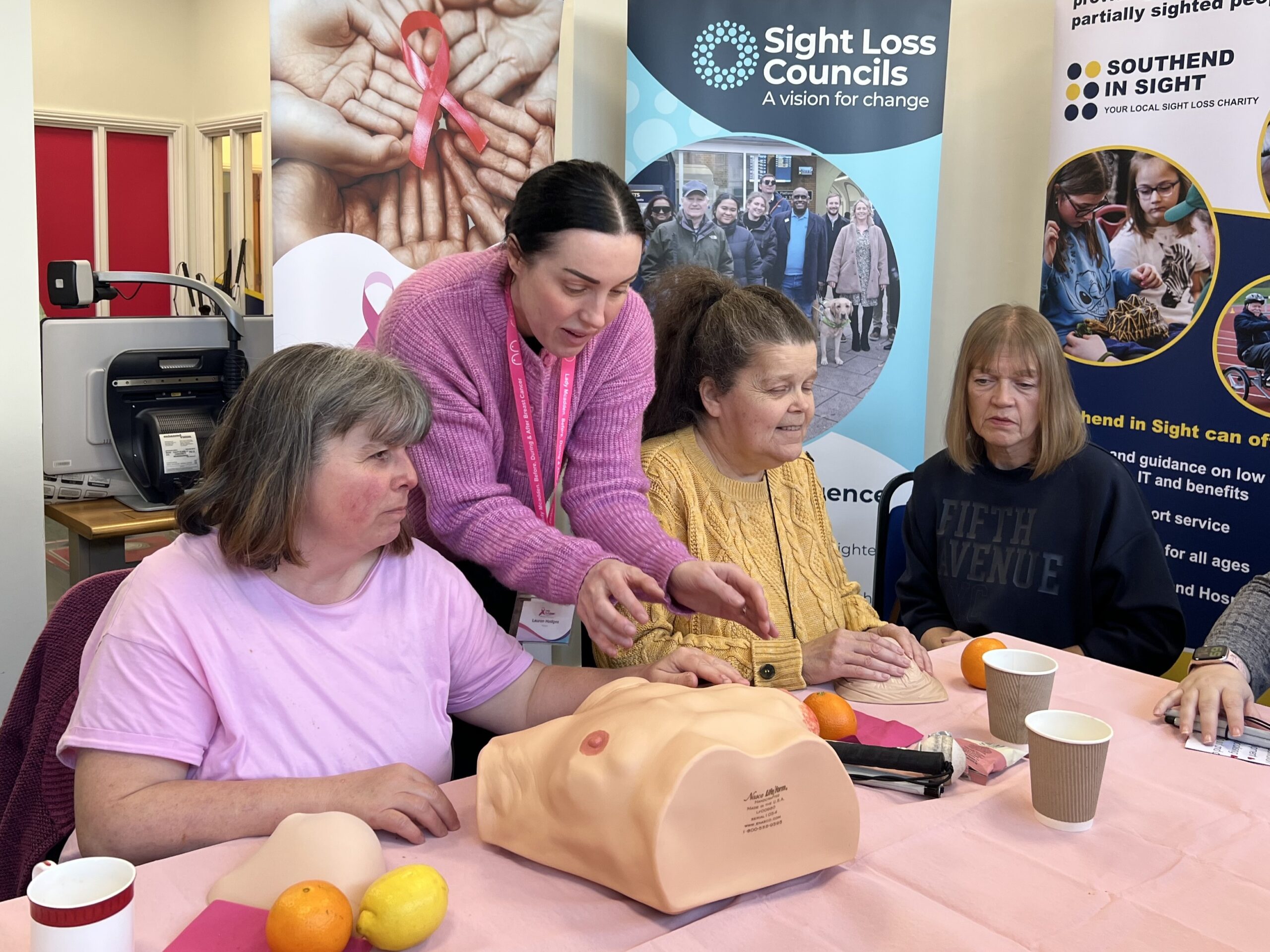 Lauren from Lady McAdden stands at a table, demonstrating how to check a silicone breast model used for breast cancer awareness. Several attendees are seated around the table, following along. Sam, Essex SLC member, is placing a hand on the model used as a tactile aid to explain changes in skin texture to be aware of.