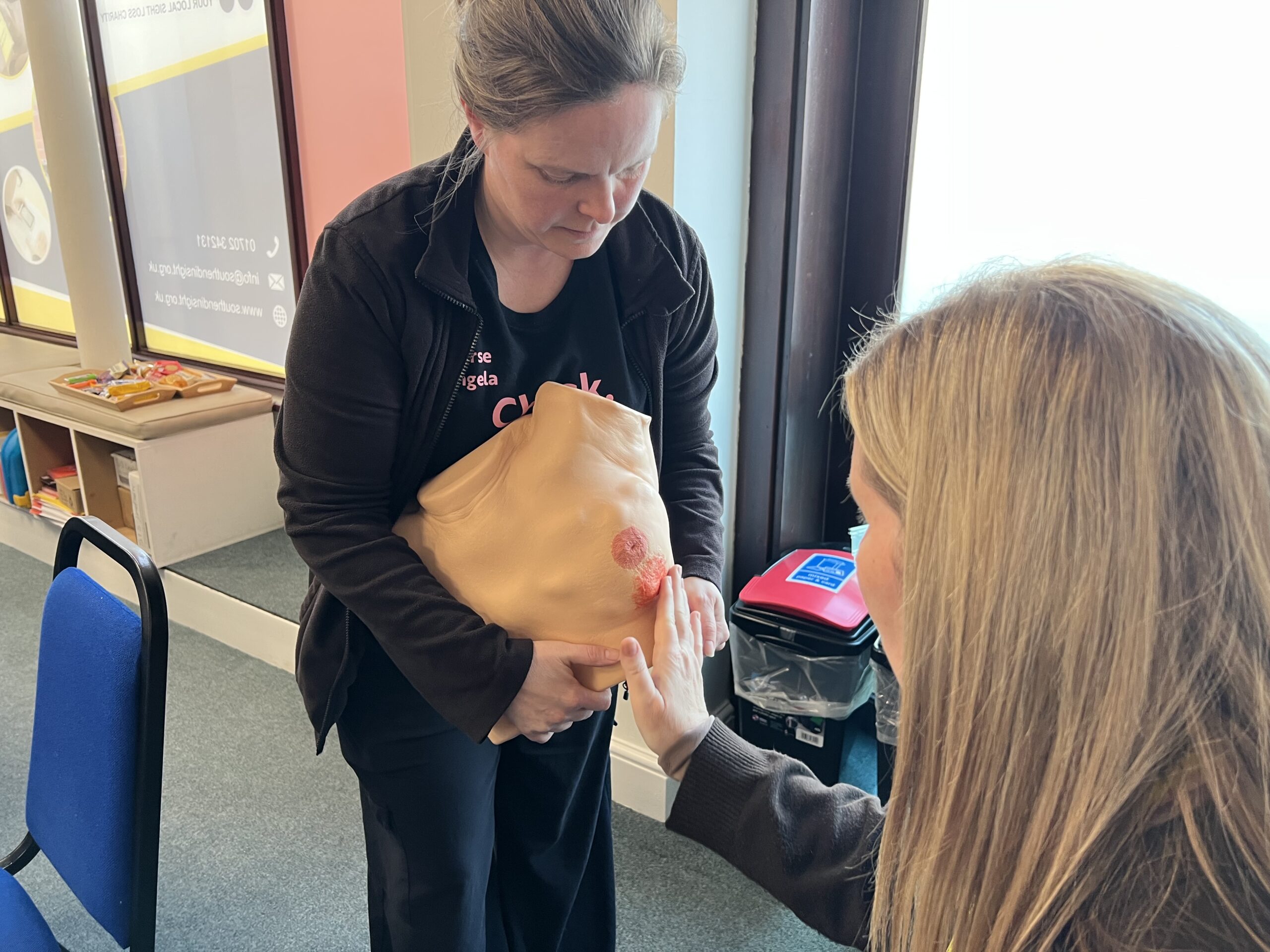 Angela from Lady McAdden is standing, holding a large silicone breast model with visible textured areas used to describe possible breast cancer symptoms. Lucy from Southend in Sight is seated in front of the model, reaching out to feel it as part of the breast cancer screening event.